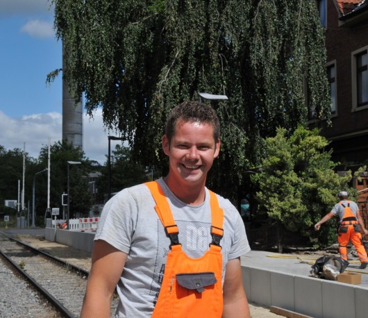 Das Bild zeigt einen lächelnden Bauarbeiter in grauem T-Shirt und orangefarbener Latzhose an einer Bahnstrecke. Im Hintergrund arbeitet ein weiterer Bauarbeiter an der Baustelle, umgeben von Bäumen und Gebäuden.