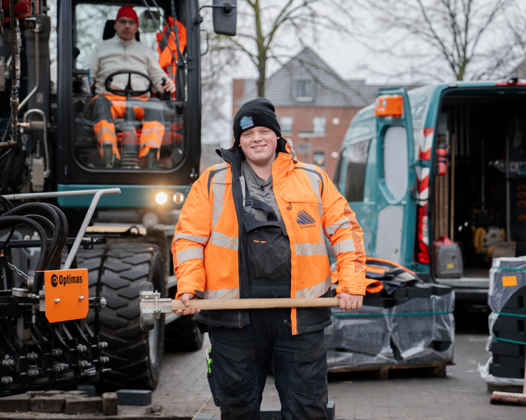 Das bild zeigt einen Bauarbeiter in orangefarbener Schutzkleidung, der lächelnd ein Werkzeug hält. Im Hintergrund ist ein Bagger mit einem weiteren Arbeiter sowie Baumaterialien und ein Firmenfahrzeug zu sehen