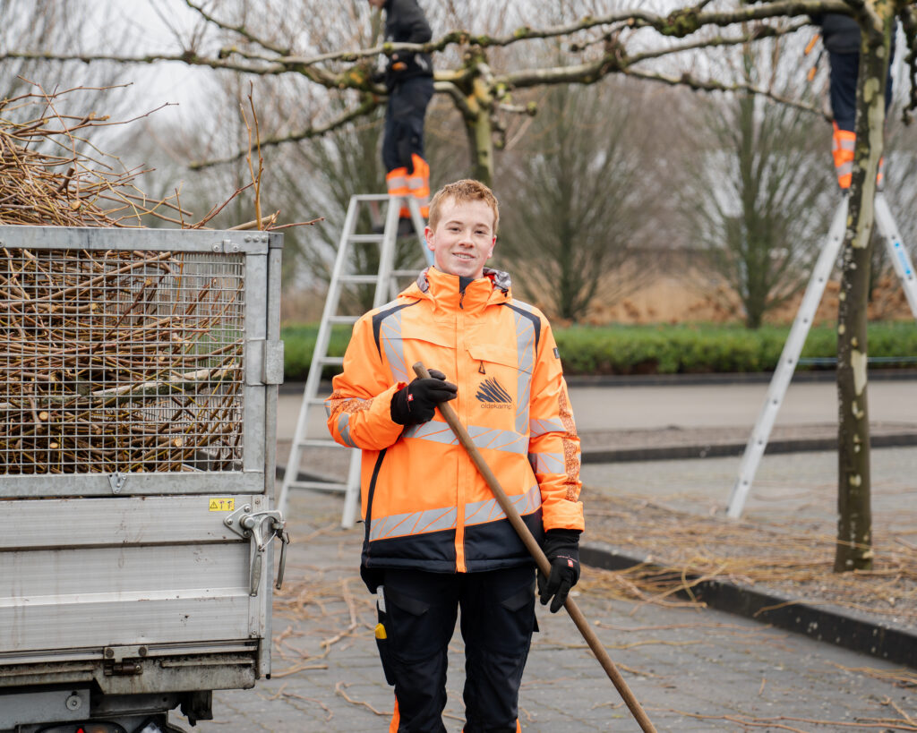 Auf dem Bild sieht man einen Arbeiter, der eine orangene Jacke anhat und einen Besen in der Hand hält. Hinter ihm sieht man ein Fahrzeug, wo die ganzen Abgeschnittenen Äste gesammelt werden